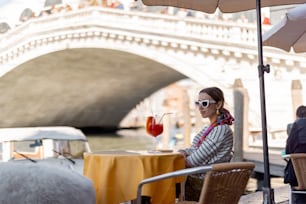 Mulher jovem desfrutando de bebida de coquetel enquanto está sentada no café ao ar livre no fundo da famosa ponte de Rialto e do Grande Canal em Veneza. Feliz tempo de lazer e férias viajando Itália