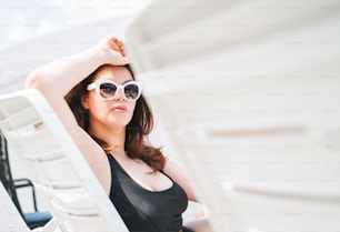 세련 된 행복 한 젊은 여성 플러스 사이즈 바디 긍정적 검은 수영복과 선글라스는 수영장 근처의 해변 안락 의자에서 인생을 즐겁게, 여름 휴가