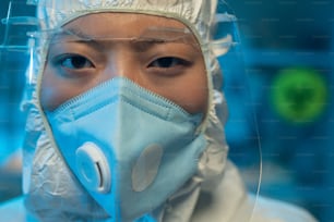 Asian female chemist or healthcare worker in white biohazard suit, respirator and protective facial screen looking at camera