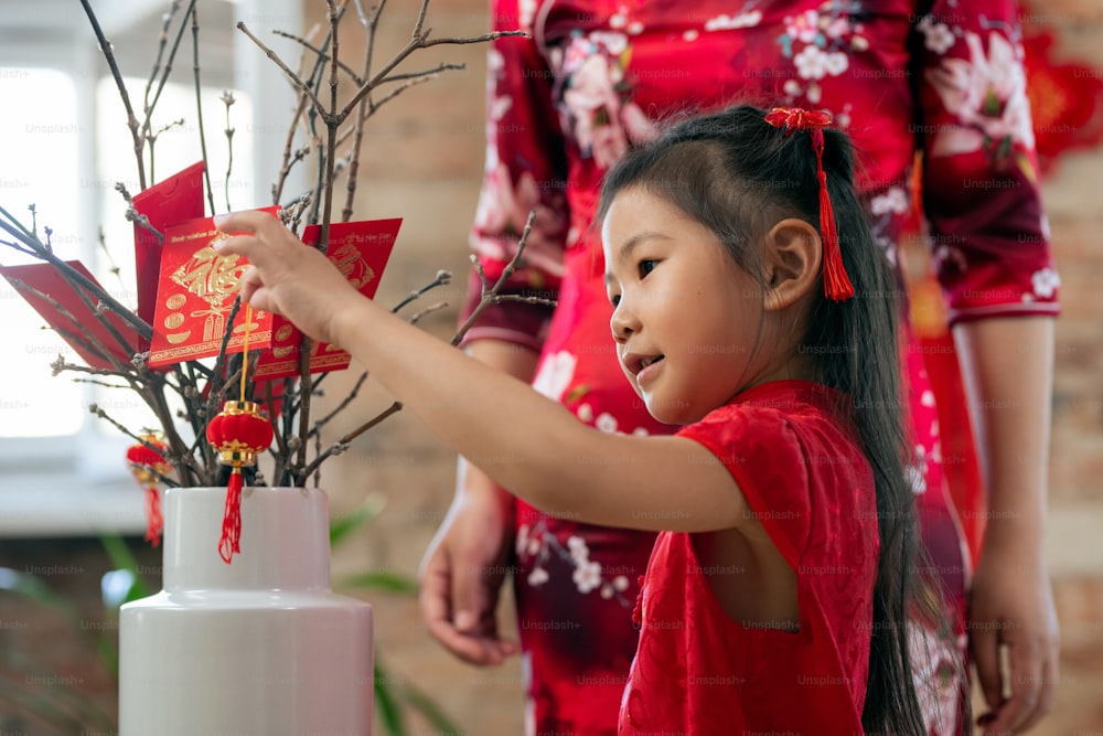 Cute little girl putting decorative lantern on branches decorated with handmade postcards saying Happy new year