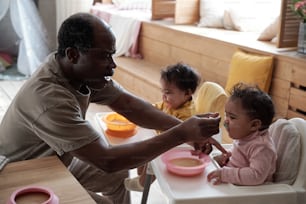 Modern mature African American man taking care of his baby daughters feeding them with fruit puree