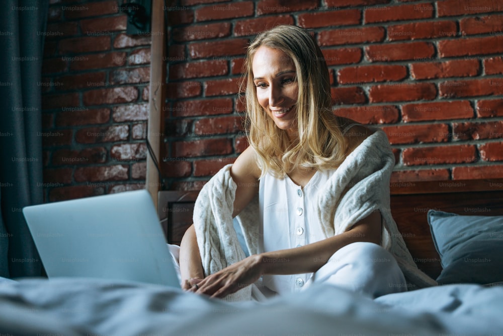 Mujer adulta de cuarenta años freelancer con cabello largo rubio en ropa casual usando computadora portátil sentada en la cama en casa