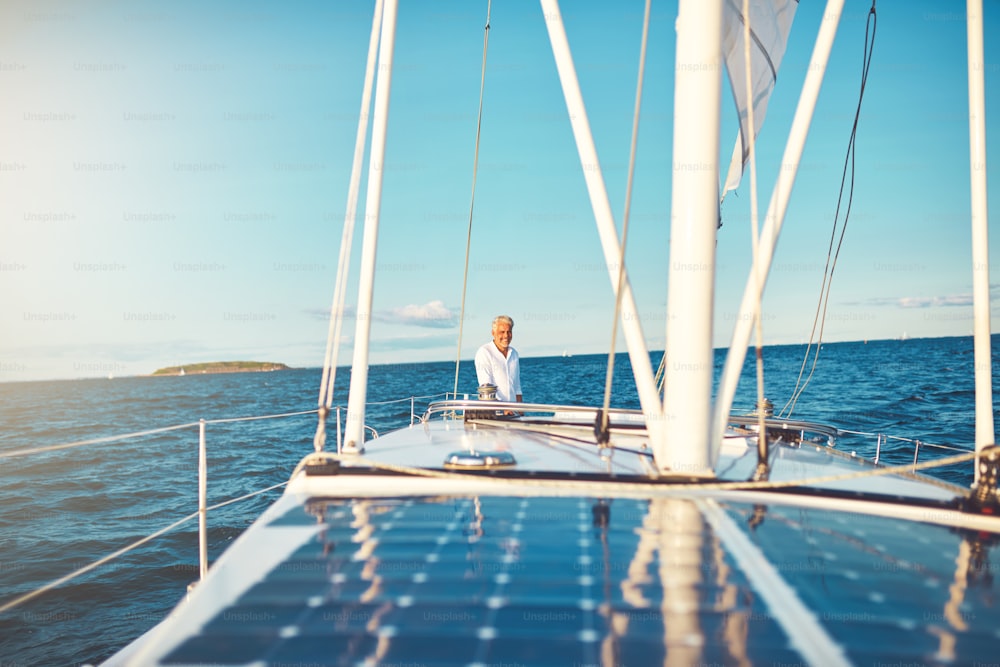 Lächelnder reifer Mann steht auf dem hinteren Deck seines Bootes, während er an einem sonnigen Tag entlang der Küste segelt