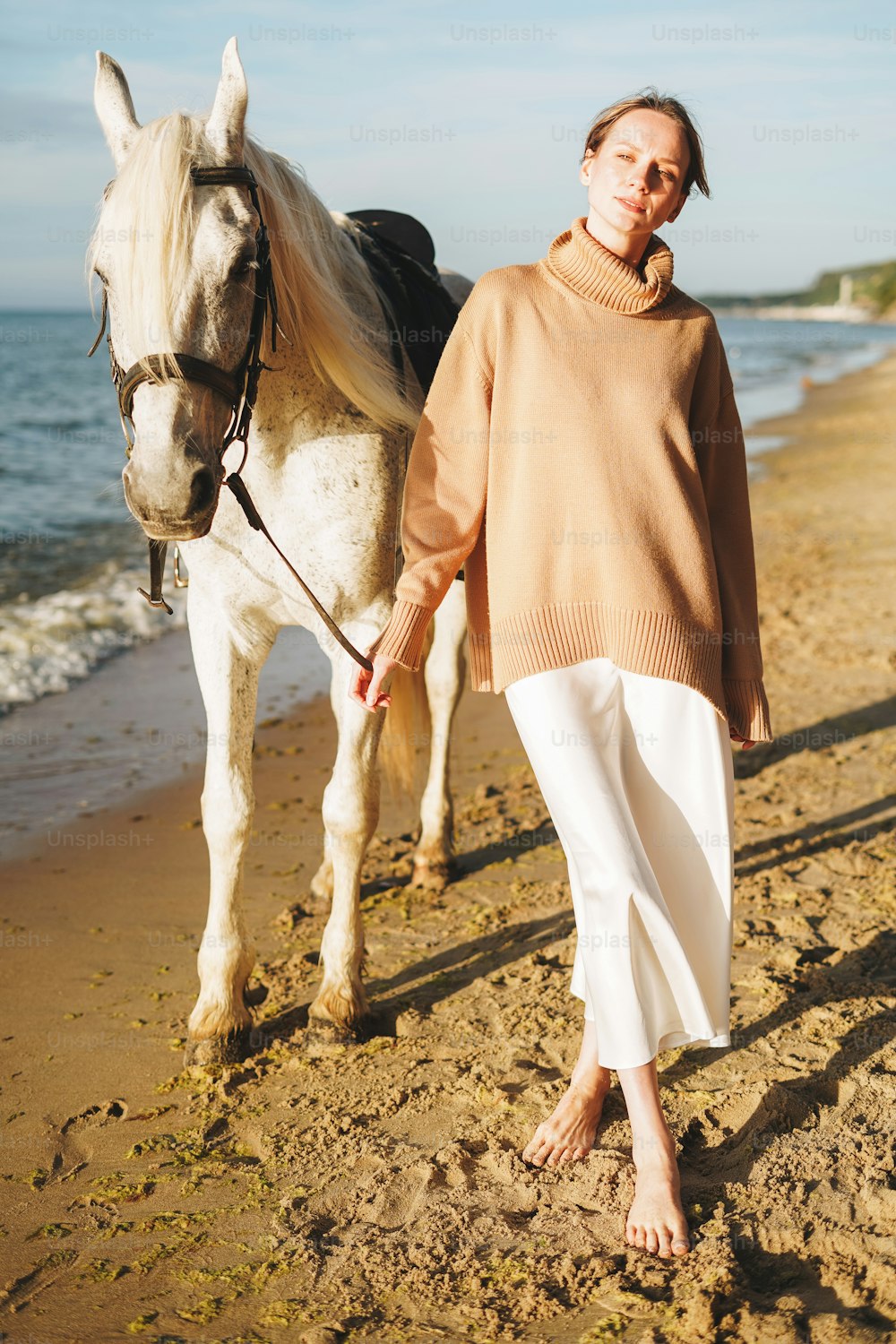 Jeune femme en pull beige avec cheval blanc sur fond de paysage marin