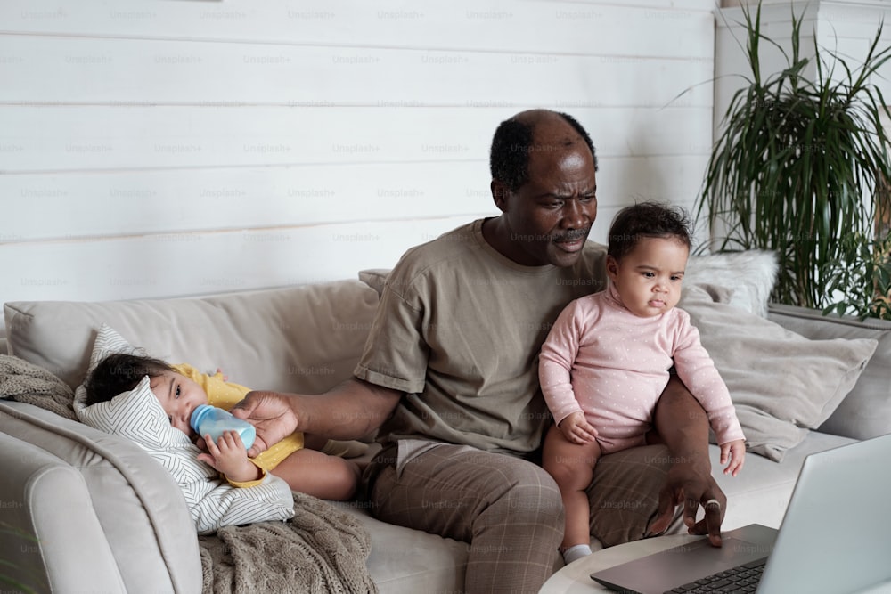 Mature African American man sitting on sofa with baby on his lap watching something on laptop while feeding another baby with formula