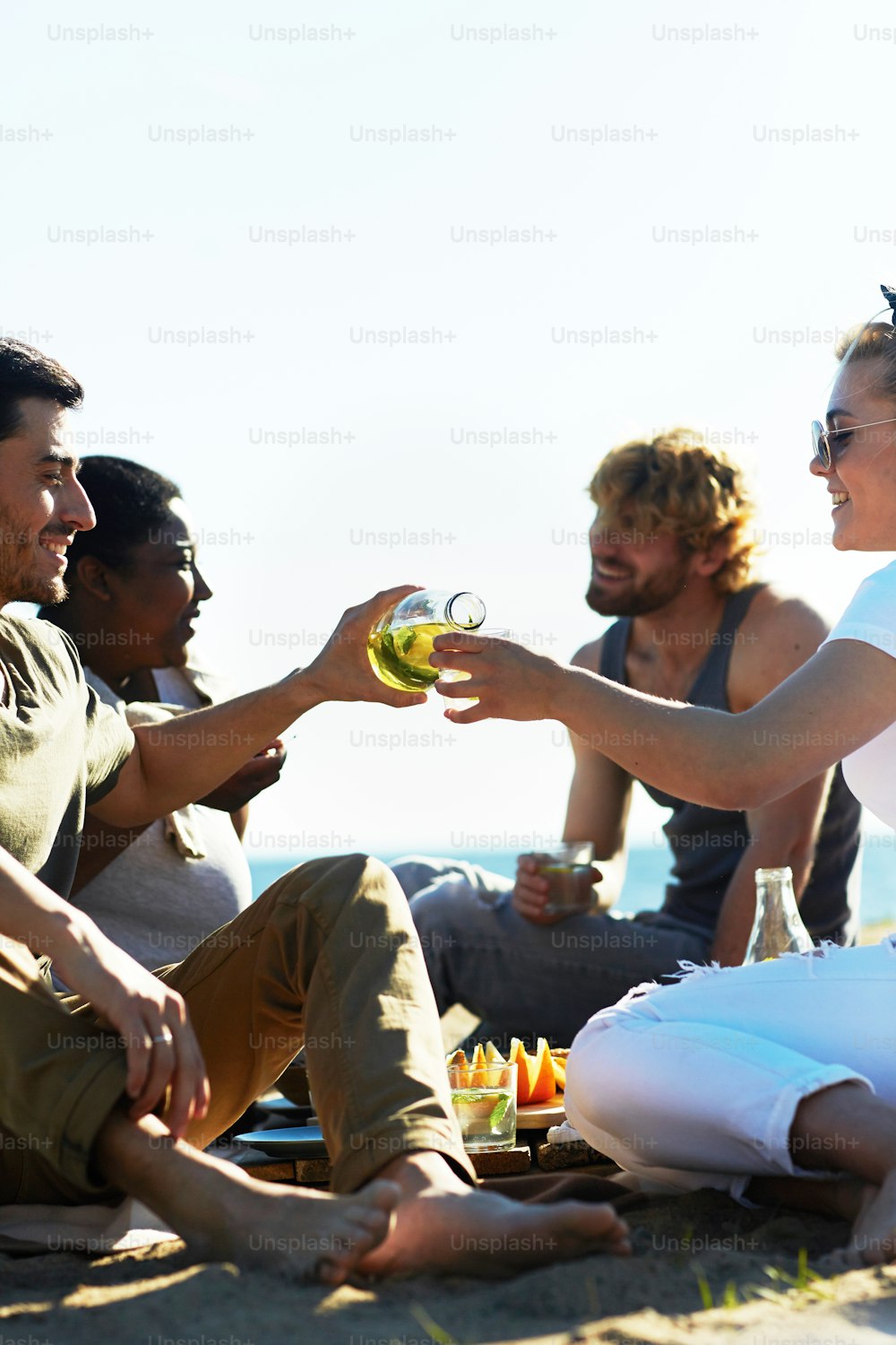 One of guys pouring lemonade from bottle to glass of girl during picnic by seaside