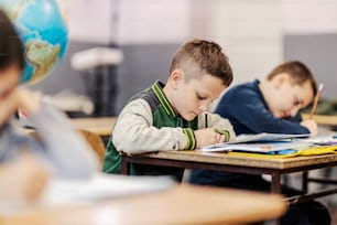 A pupil writes schoolwork while sitting at elementary school with classmates.