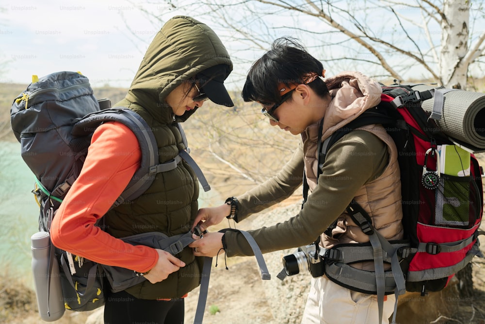 Vista lateral da jovem mulher asiática caminhando junto com a melhor amiga ou irmã ajudando-a a ajustar e consertar mochila de viagem pesada