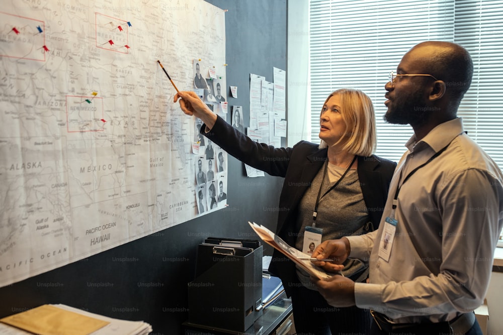 Two FBI agents looking at map on board while mature female pointing at it and making suggestions about possible location of criminal