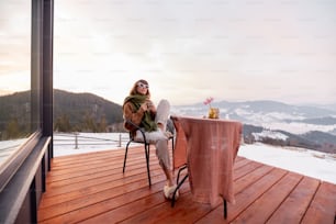 Frau sitzt im Winter auf der Terrasse mit herrlichem Blick auf die Berge, genießt Getränke und wunderschöne Landschaft bei Sonnenaufgang. Konzept der Ruhe und Flucht in die Natur