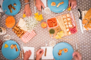 4人の年配の男女の昼食中のテーブルの高い眺め。モルタデッラ、ニンジン、サラミ、パンなどの食べ物が入った明るい画像。テーブルの上にはたくさんの手が食べようと待っています