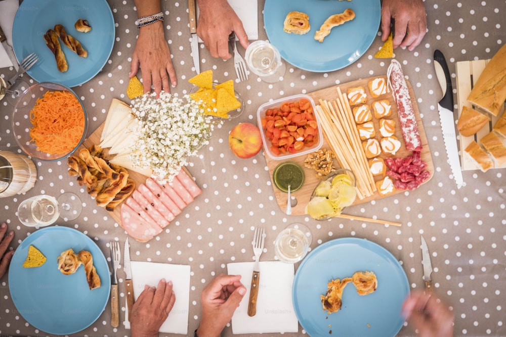 high view of a table during a lunch for 4 senior men and women. bright image with food like mortadella, carrots, salami, bread and more. many hands on the table waiting to eat