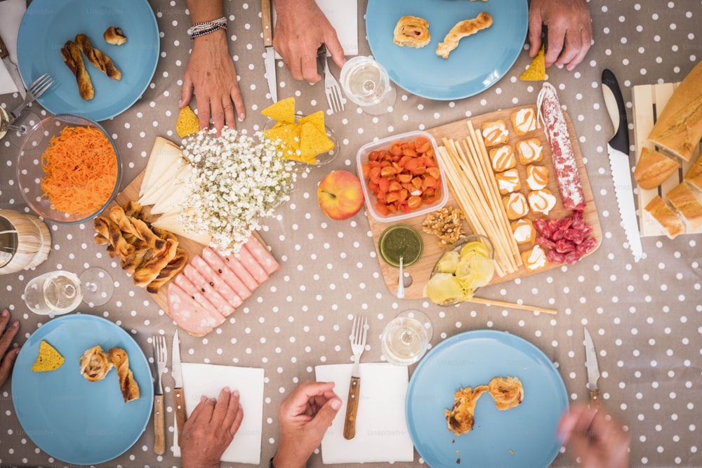 Vista alta de una mesa durante un almuerzo para 4 hombres y mujeres mayores. Imagen brillante con alimentos como mortadela, zanahorias, salami, pan y más. muchas manos en la mesa esperando para comer