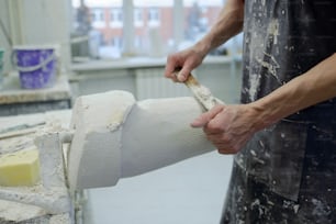 Hands of young prosthetic factory worker in apron polishing plaster cast with chisel while standing by workplace in front of camera