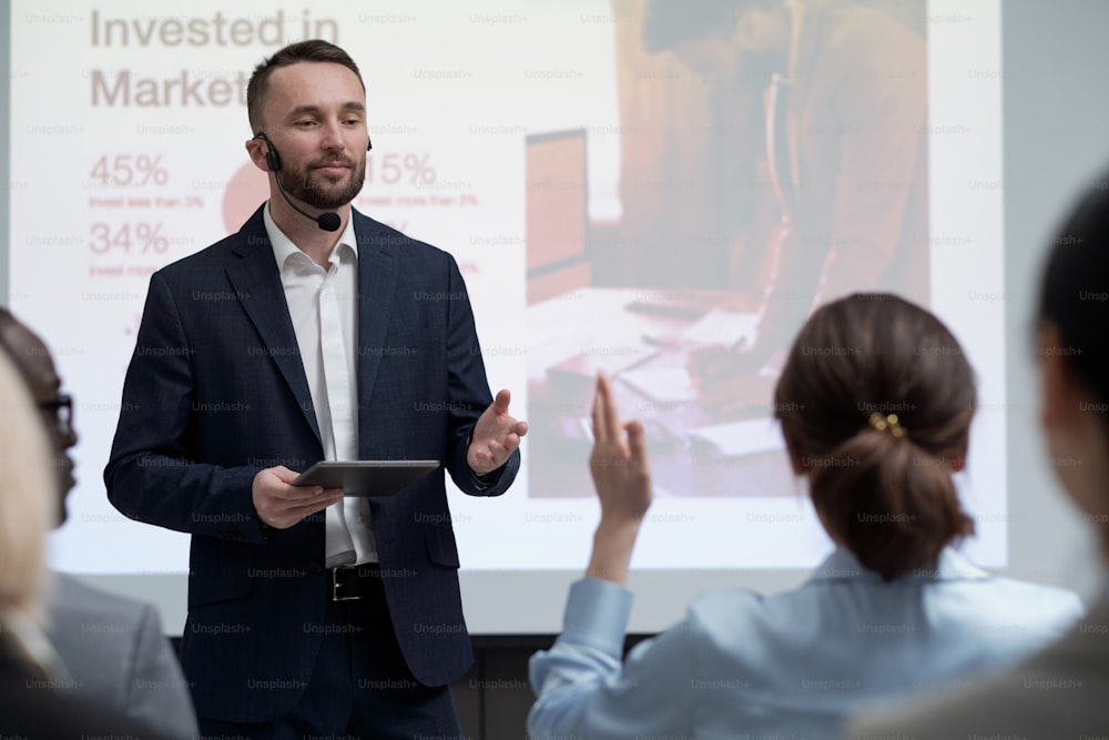 Young contemporary businessman pointing at one of colleagues during presentation of revenue invested in marketing at conference