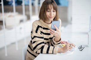 Young woman using phone while sitting at restaurant and having lunch with a sushi rolls. Caucasian woman wearing striped sweater sitting at modern white restaurant