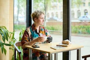 Mujer joven serena con discapacidad física mirando a través de una ventana grande mientras está sentada junto a la mesa en la cafetería y tomando café con la merienda