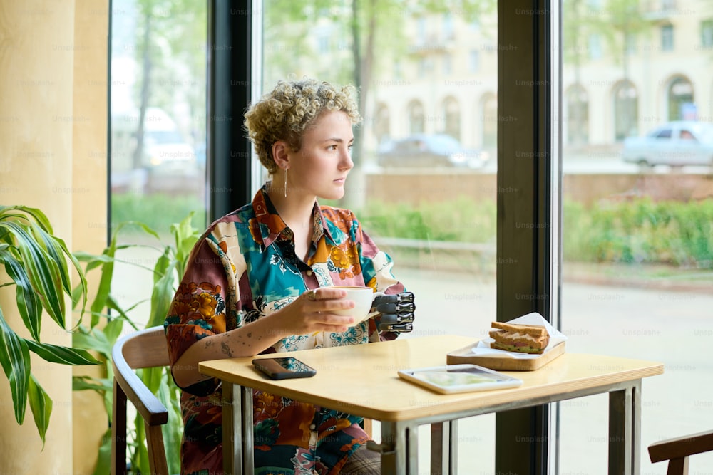 Junge ruhige Frau mit körperlicher Beeinträchtigung, die durch ein großes Fenster schaut, während sie am Tisch im Café sitzt und Kaffee mit Snack trinkt