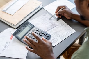 Manos de hombre africano presionando botones de calculadora mientras verifica datos en papeles financieros y facturas impagas por tabla