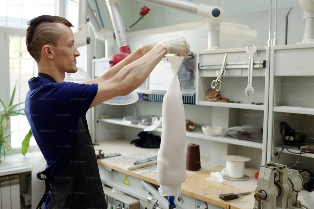 Giovane uomo in guanti e grembiule che mette un tessuto speciale sull'arto artificiale mentre forma la forma dell'incavo protesico in woekshop