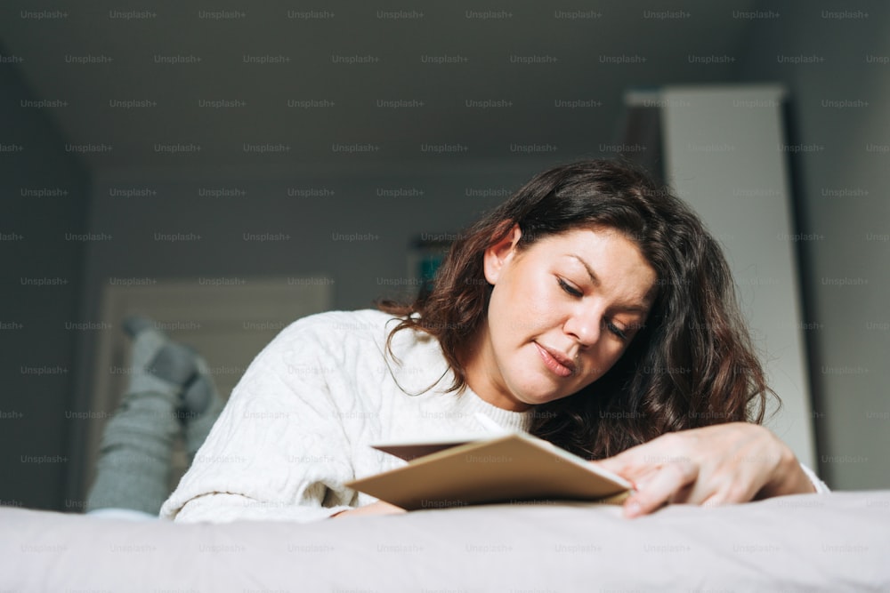 Giovane donna bruna adulta di quarant'anni plus size body positive in maglione lavorato a maglia bianco scrive note in agenda sul letto di casa