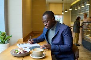 Hombre de negocios serio africano que usa una tableta y llena el documento en la mesa de la cafetería durante su desayuno