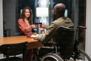 Happy young interracial couple sitting by festive table and toasting with red wine during romantic dinner at home or restaurant