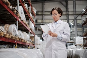 Portrait of female factory worker wearing glasses and white robe, using tablet for quality control and logistic purposes at polymer plastic manufacturing, standing between shelves with polymer rolls