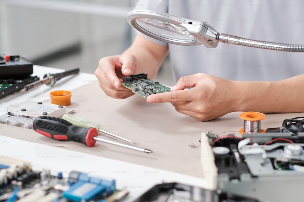 Hands of repairperson holding circuit boad of electronic device behind magnifying glass