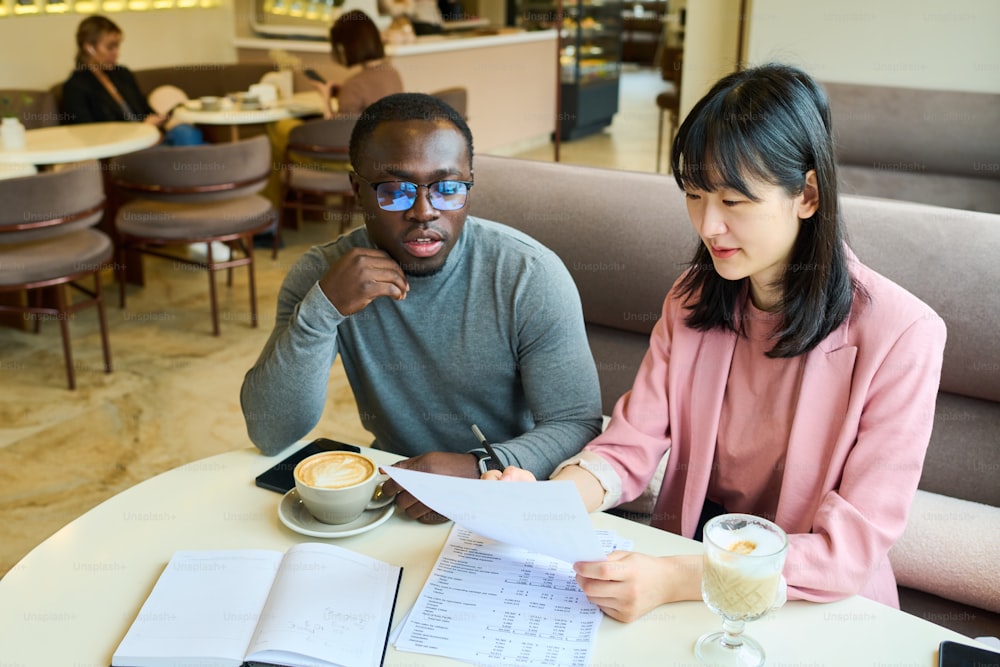 Business colleagues sitting at table with coffee drinks and discussing documents during meeting at cafe