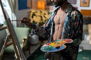 컬러 팔레트를 가진 젊은 섹시한 남자의 중간 부분은 이젤 앞에 서 있는 동안 캔버스에 새로운 그림을 만듭니다.