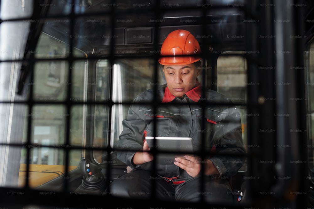 Young serious multi-ethnic female with tablet looking for technical data in the internet while sitting inside construction machine