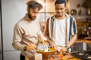 Dois caras de etnia diferente cozinhando comida vegana saudável na cozinha em casa. Conceito de amizade ou relacionamento masculino próximo como gay. Ideia de alimentação saudável