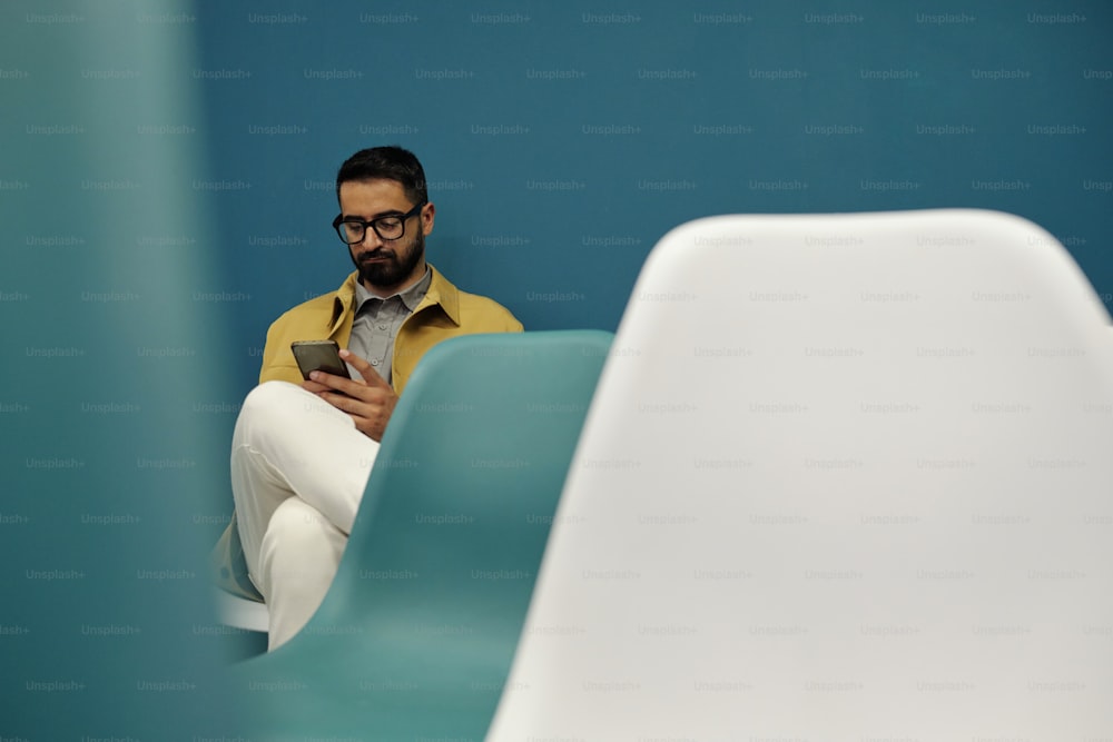Seriöser Geschäftsmann mit Smartphone, der im Hörsaal zwischen weißen und blauen Stühlen sitzt und online Daten oder Fotos durchschaut
