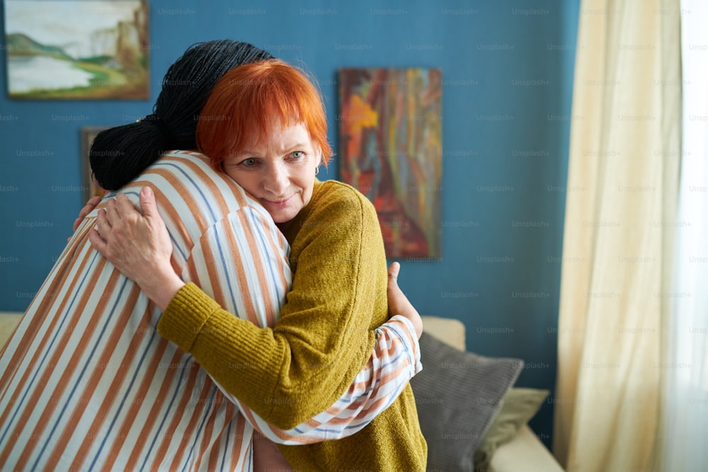 Senior woman with red hair embracing volunteer while they standing in living room at home
