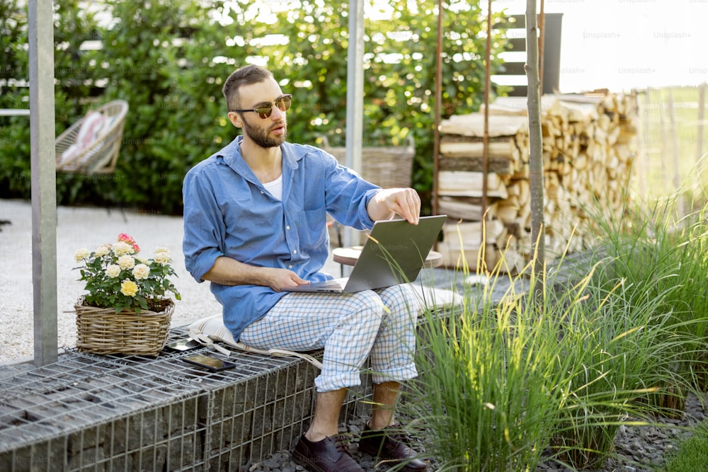 Cool guy works on laptop while sitting on porch of his country house. Concept of remote work at cozy home workplace and freelance