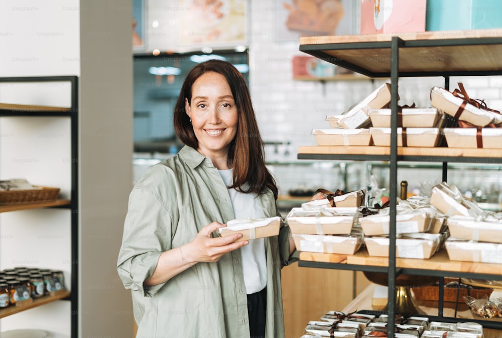 Adult smiling brunette woman forty years with long hair in stylish shirt choosing sweets in cafe, woman having shopping in confectionery