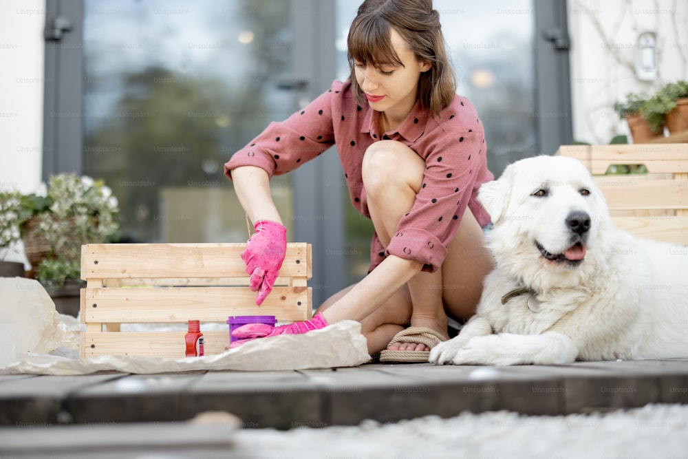 Young woman painting wooden box in pink color, doing housework while sitting with her dog on terrace of her house. DIY concept. Idea of friendship with pets