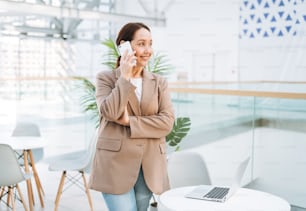 Adult smiling brunette business woman forty years with long hair in stylish beige suit and jeans working on laptop at public place, open space office