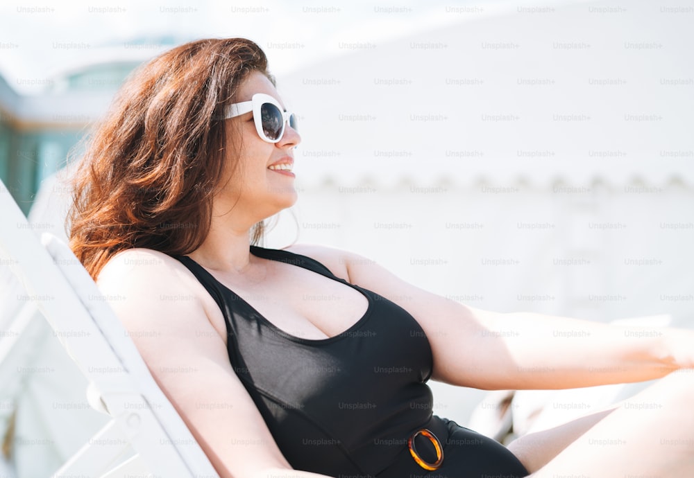 Stylish happy young woman plus size body positive in black swimsuit and sunglasses enjoing life rest on beach lounger near the pool, summer vacations