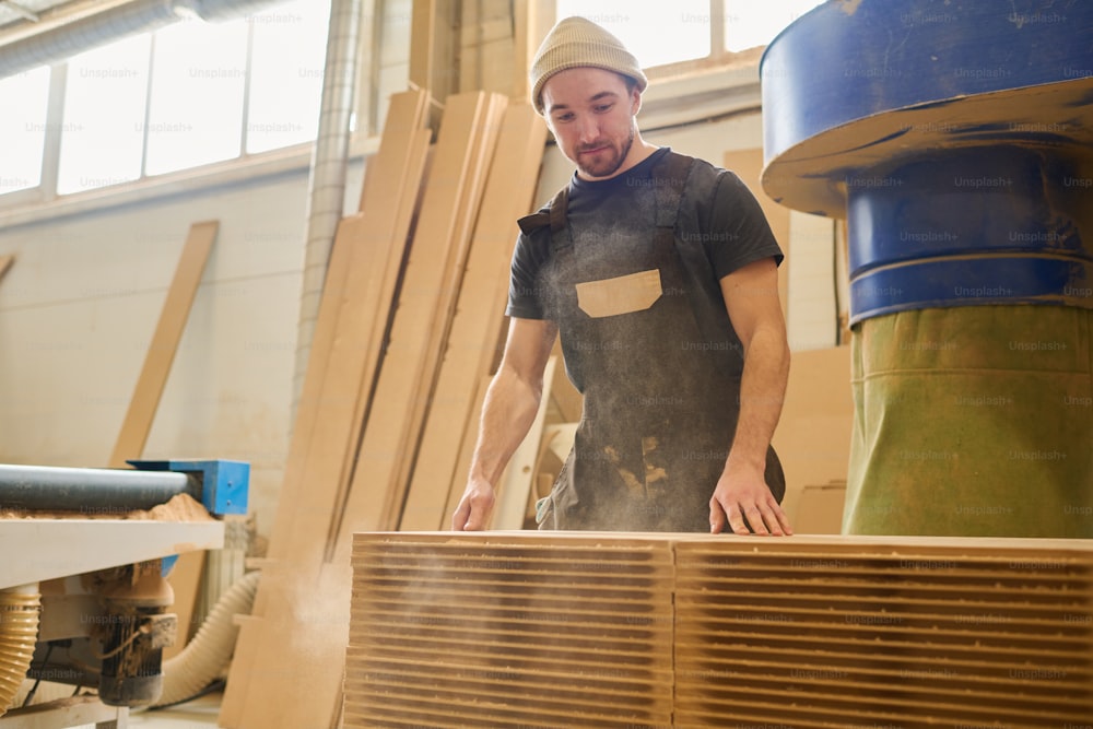 Jeune ouvrier en uniforme debout sur une pile avec de fines planches de bois pendant son travail à l’usine de meubles