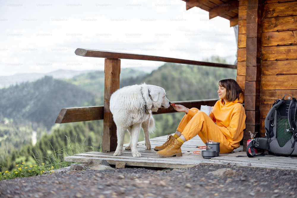 若い女性は素晴らしい山の風景を楽しみ、ハイキングのために昇華した食べ物を食べながら、自然の家の木製のテラスに犬と一緒に座っています。自然の中での孤独とペットとの旅行の概念
