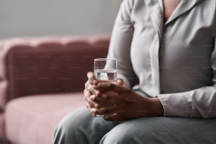 Hands of young worried female patient of counselor holding glass of water while sitting on couch and describing her problems