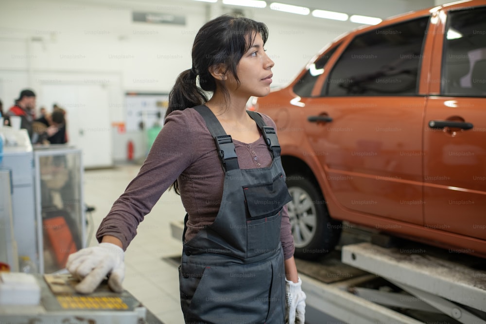 Jeune femme hispanique confiante en salopette et gants travaillant dans le garage d’un service d’entretien automobile contemporain