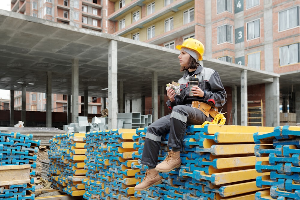 Young female worker of construction site holding sandwich and mug with hot tea while sitting on stack of building materials at lunch break