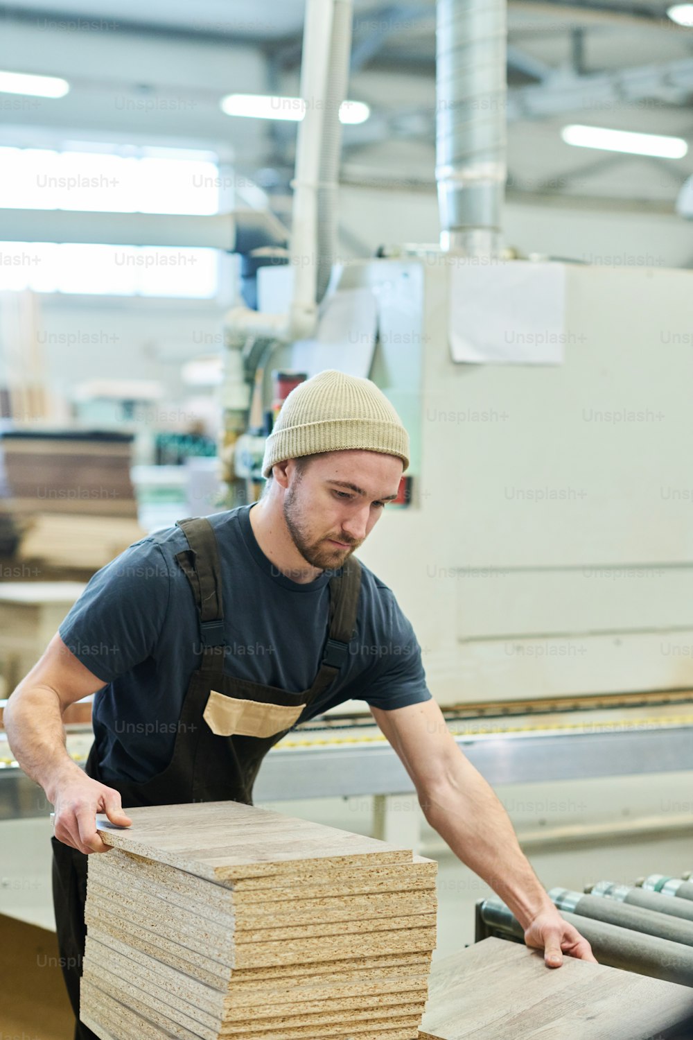 Young carpenter in uniform working with wooden boards at machine at furniture factory