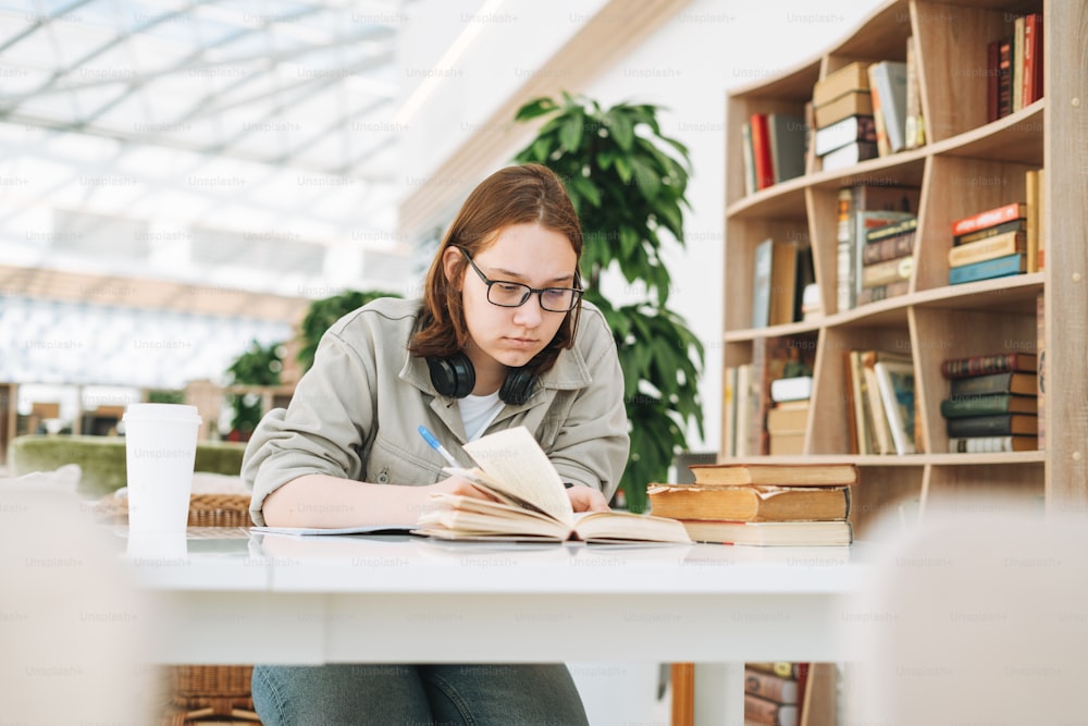 Jovem morena adolescente estudante universitária em óculos fazendo lição de casa com livros no local público da biblioteca moderna verde