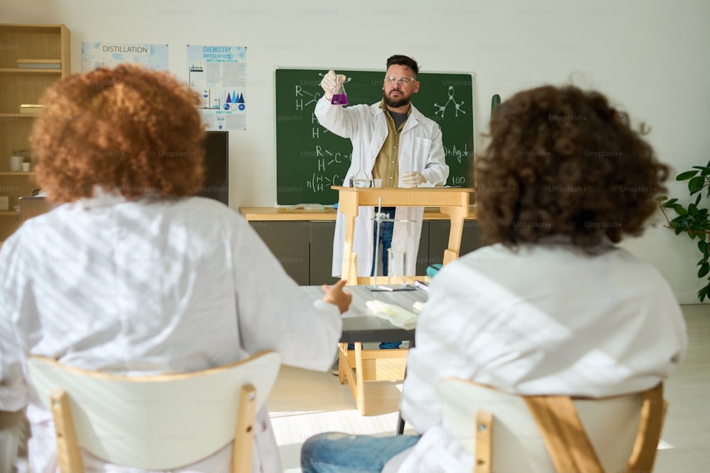 두 가지 다양한 유체를 혼합하여 얻은 액체 물질로 튜브를 학생들에게 보여주는 실험실 코트의 자신감 있는 화학 교사