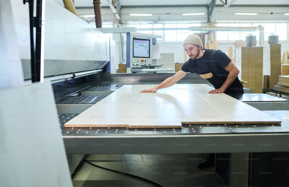 Giovane operaio in uniforme che mette tavole di legno su attrezzature moderne durante il lavoro in fabbrica di mobili