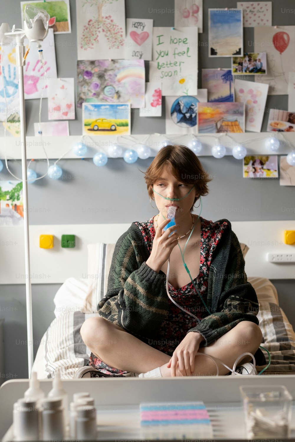 Adolescente enferma con tubo nasal de oxígeno respirando por la boca con inhalador mientras está sentada en la cama en la sala del hospital
